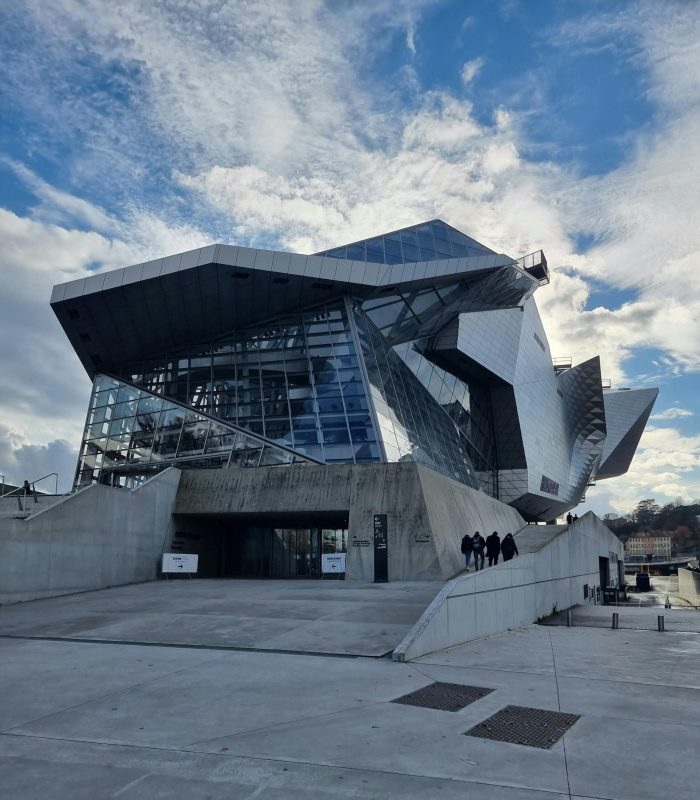 Un bâtiment moderne et anguleux avec des façades en verre et en métal sous un ciel partiellement nuageux. Plusieurs personnes se dirigent vers l'entrée en empruntant une large rampe en béton.