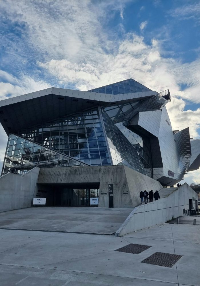 Un bâtiment moderne et anguleux avec des façades en verre et en métal sous un ciel partiellement nuageux. Plusieurs personnes se dirigent vers l'entrée en empruntant une large rampe en béton.