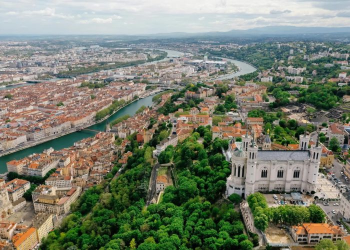 Aerial view of Lyon, France, with the Notre-Dame de Fourvière basilica surrounded by lush vegetation, the city's red-roofed buildings and the Saône and Rhône rivers winding through the landscape.