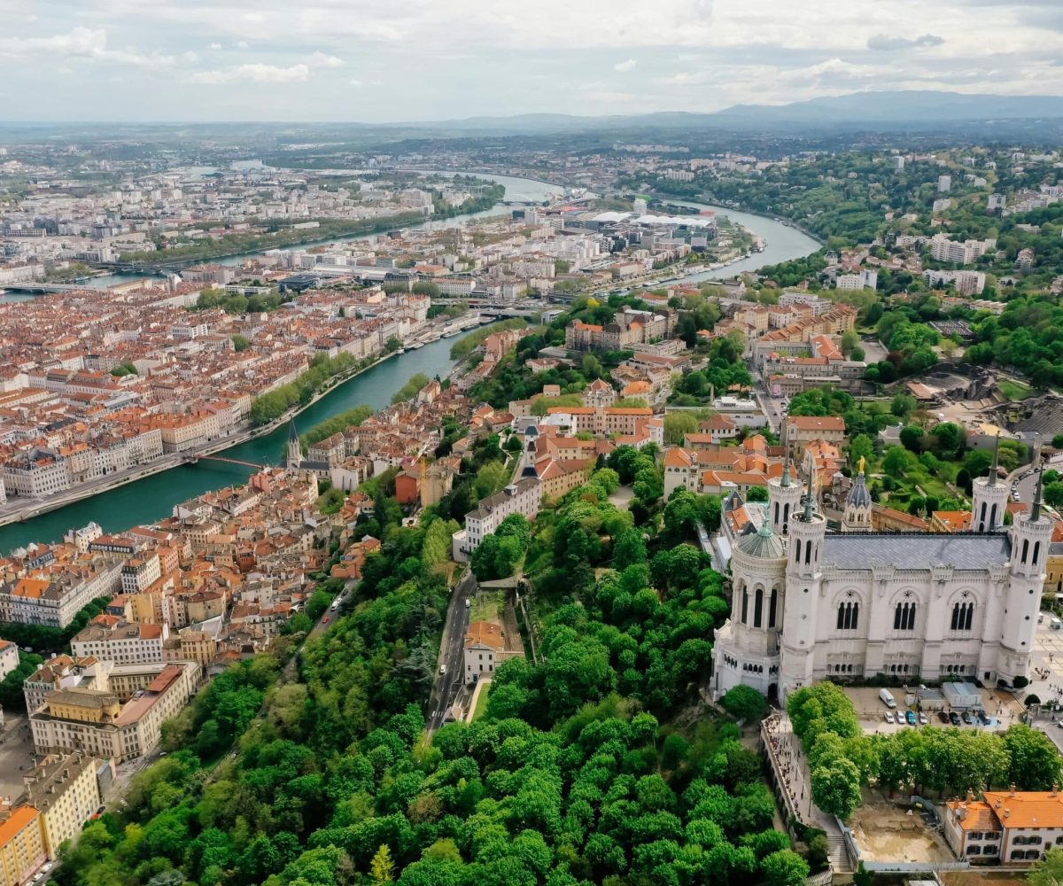 Vue aérienne de Lyon, France, avec la basilique Notre-Dame de Fourvière entourée d'une végétation luxuriante, les bâtiments à toits rouges de la ville et la Saône et le Rhône serpentant dans le paysage.