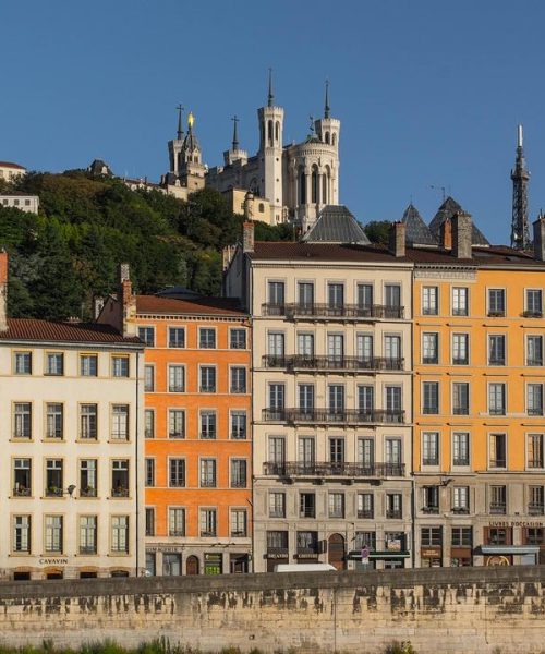 Des bâtiments colorés bordent un talus en pierre à Lyon, en France. La basilique Notre-Dame de Fourvière, située au sommet d'une colline, et une verdure luxuriante sont visibles à l'arrière-plan, sous un ciel bleu clair.
