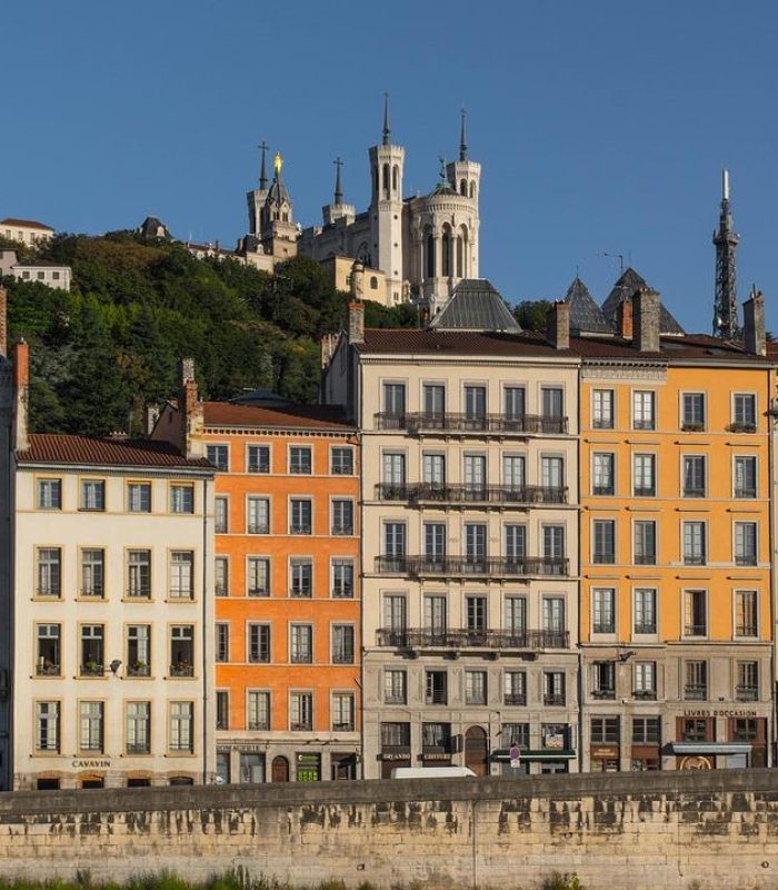 Des bâtiments colorés bordent un talus en pierre à Lyon, en France. La basilique Notre-Dame de Fourvière, située au sommet d'une colline, et une verdure luxuriante sont visibles à l'arrière-plan, sous un ciel bleu clair.