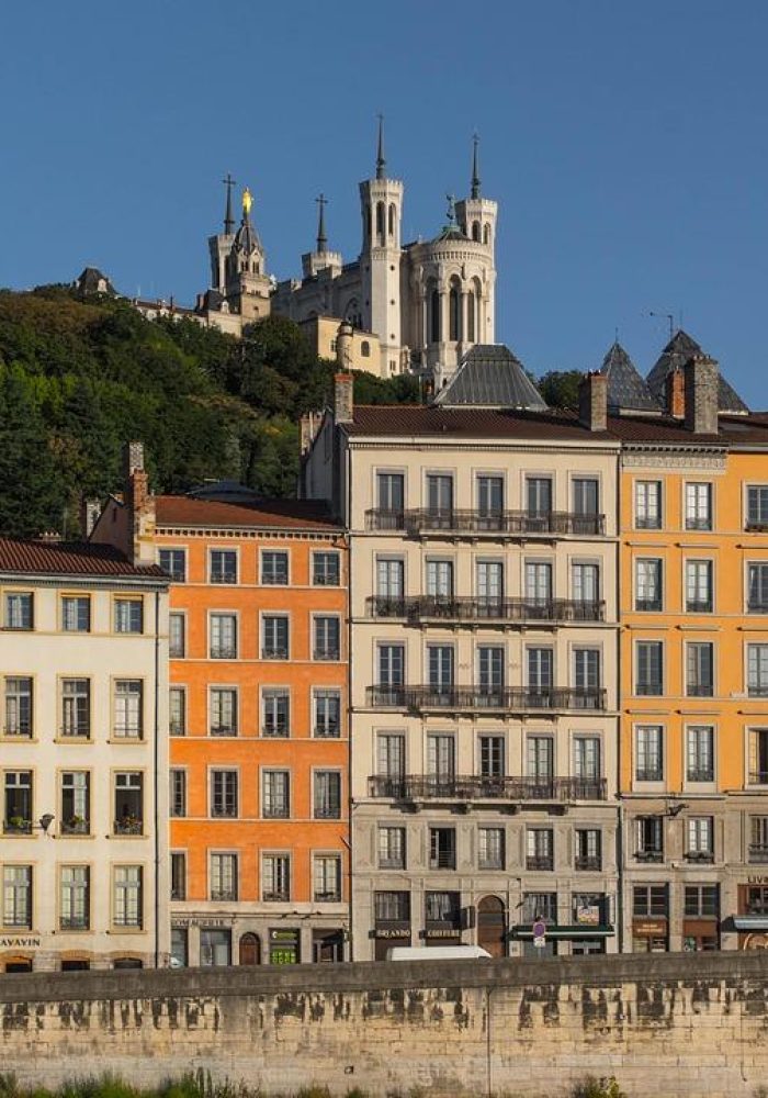 Des bâtiments colorés bordent un talus en pierre à Lyon, en France. La basilique Notre-Dame de Fourvière, située au sommet d'une colline, et une verdure luxuriante sont visibles à l'arrière-plan, sous un ciel bleu clair.