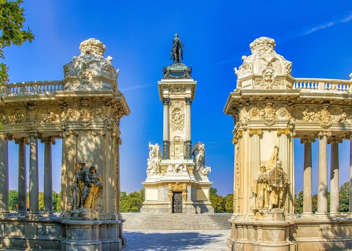 Un grand monument avec une haute colonne centrale surmontée d'une statue d'homme, flanquée de colonnades ornées et de sculptures, le tout dans un ciel bleu clair dans un parc.