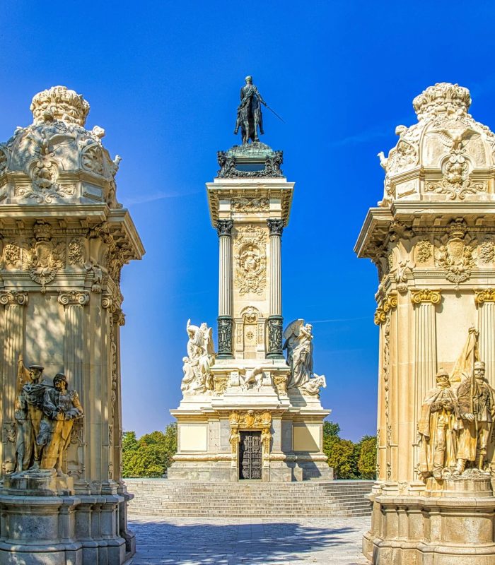 Un grand monument avec une haute colonne centrale surmontée d'une statue d'homme, flanquée de colonnades ornées et de sculptures, le tout dans un ciel bleu clair dans un parc.