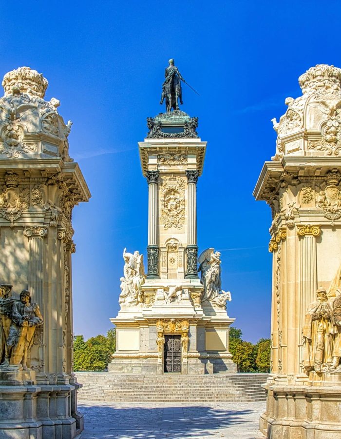 Un grand monument avec une haute colonne centrale surmontée d'une statue d'homme, flanquée de colonnades ornées et de sculptures, le tout dans un ciel bleu clair dans un parc.