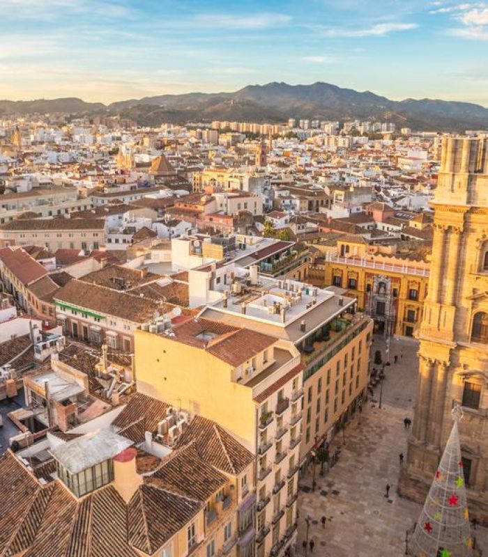 Vue aérienne d'une ville éclairée par le soleil, avec une cathédrale historique bien en vue, des rues étroites et des bâtiments densément peuplés, avec pour toile de fond des montagnes lointaines sous un ciel bleu.