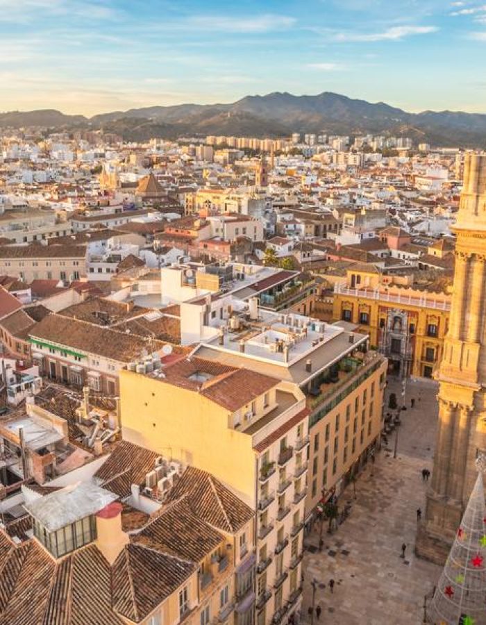 Vue aérienne d'une ville éclairée par le soleil, avec une cathédrale historique bien en vue, des rues étroites et des bâtiments densément peuplés, avec pour toile de fond des montagnes lointaines sous un ciel bleu.
