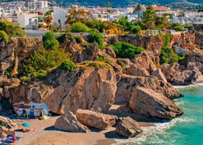 Des parasols et des transats colorés bordent un rivage sablonneux à l'eau turquoise, avec des falaises rocheuses et des bâtiments blancs au-dessus, entourés d'une verdure luxuriante et d'une montagne lointaine en toile de fond.