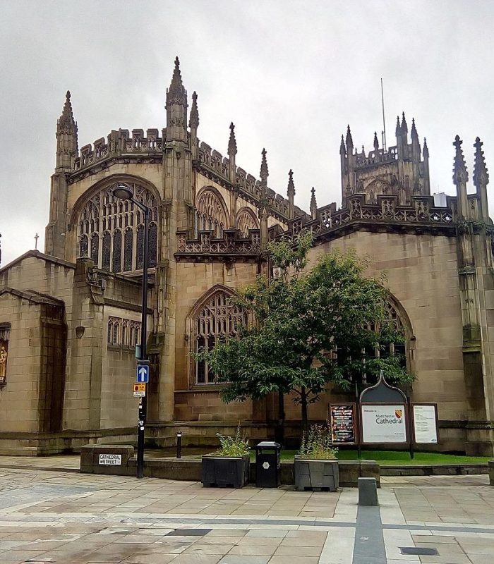 A large historic stone church, with Gothic architecture and large pointed arches, stands on an overcast day. There are road signs and trees in front, and the area is paved with stone.