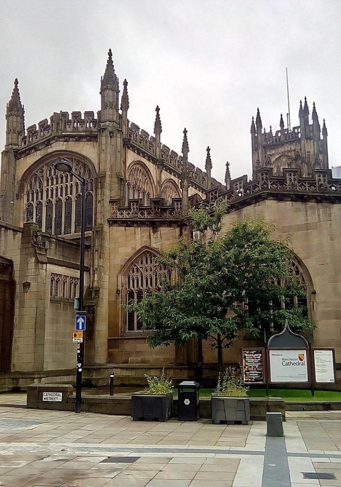 A large historic stone church, with Gothic architecture and large pointed arches, stands on an overcast day. There are road signs and trees in front, and the area is paved with stone.