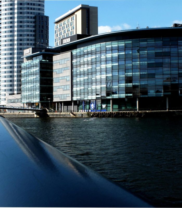 Modern glass office buildings and a large residential tower line the waterfront on a sunny day, seen from the railing of a bridge in the foreground, with blue skies and reflections on the water.