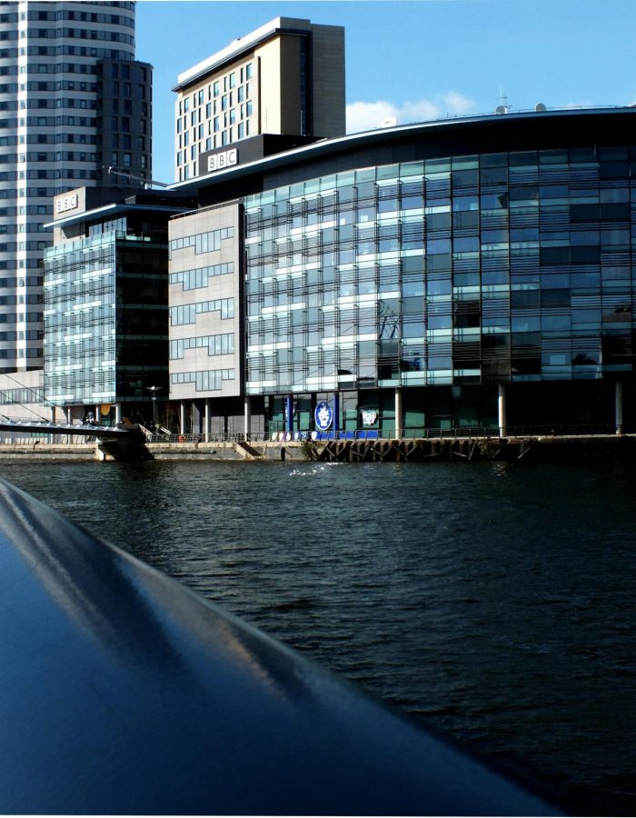 Modern glass office buildings and a large residential tower line the waterfront on a sunny day, seen from the railing of a bridge in the foreground, with blue skies and reflections on the water.