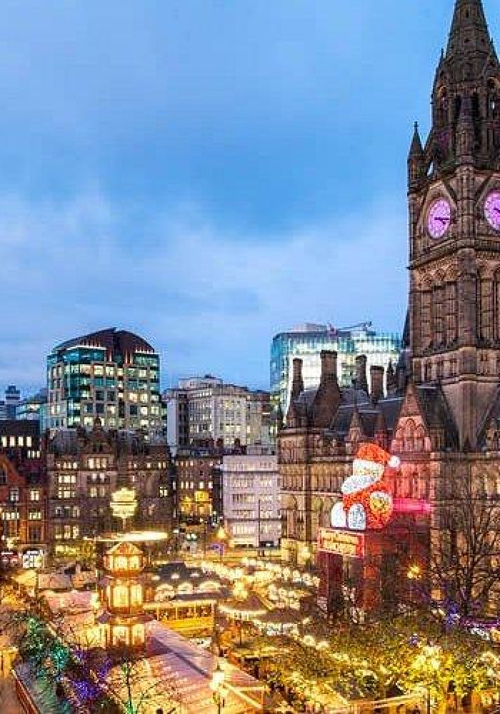 A festive city scene at dusk shows Manchester Town Hall illuminated with its clock tower, surrounded by Christmas lights, market stalls and bustling crowds in the city centre.
