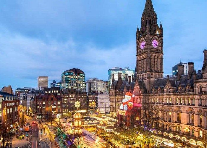 Une scène de ville festive au crépuscule montre l'hôtel de ville de Manchester illuminé avec sa tour d'horloge, entouré de lumières de Noël, d'étals de marché et d'une foule animée dans le centre ville.