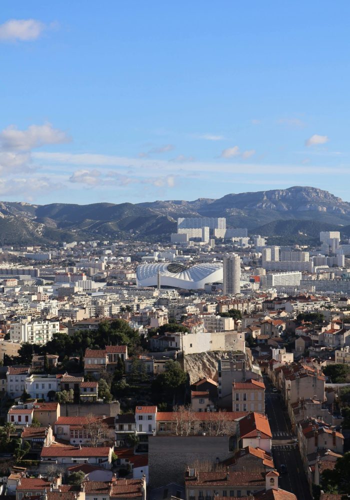 A cityscape with red-roofed buildings in the foreground, a large modern stadium in the middle, and mountains under a blue sky with scattered clouds in the background.