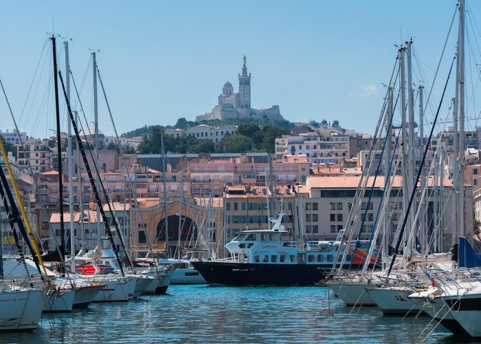 Boats and yachts are moored in a marina with urban buildings behind them; in the background, a large church stands on a hilltop under a clear blue sky.