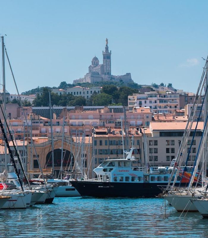 Des bateaux et des yachts sont amarrés dans un port de plaisance avec des bâtiments urbains derrière eux ; à l'arrière-plan, une grande église se dresse au sommet d'une colline sous un ciel bleu clair.