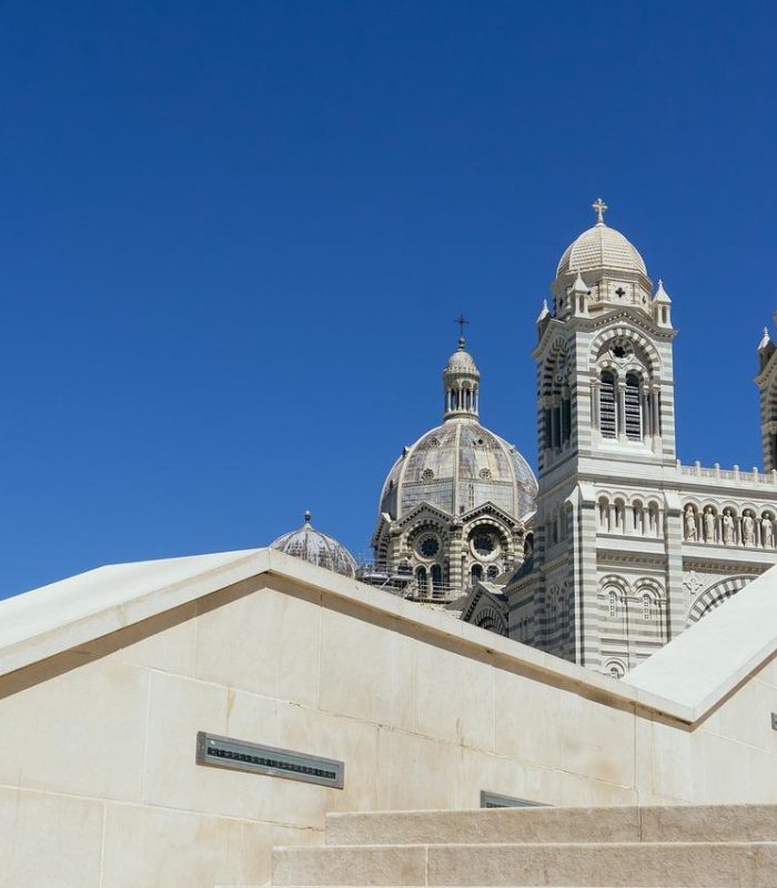 L'image montre les dômes et les tours d'une grande cathédrale derrière des marches et des murs en pierre de couleur crème, sur fond de ciel bleu clair.