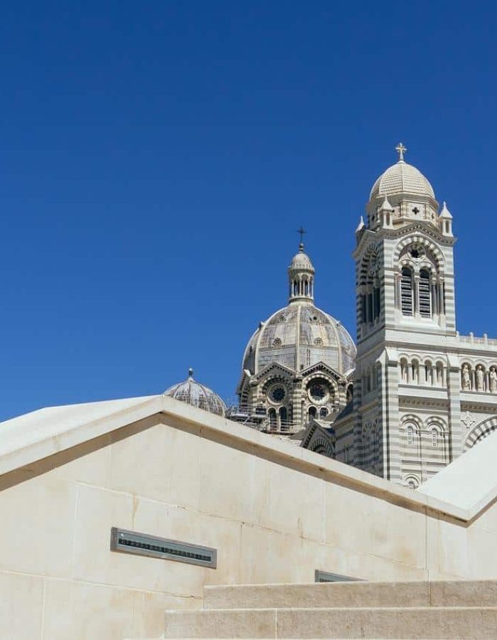 L'image montre les dômes et les tours d'une grande cathédrale derrière des marches et des murs en pierre de couleur crème, sur fond de ciel bleu clair.