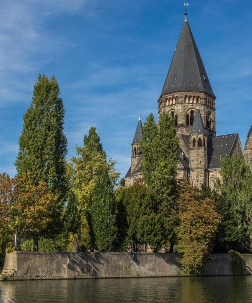 Une grande église en pierre avec une haute flèche se dresse parmi les arbres au bord d'une rivière calme sous un ciel bleu clair. Quelques bâtiments sont partiellement visibles sur la gauche.