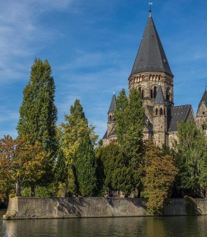 A large stone church with a high spire stands among the trees on the banks of a calm river under a clear blue sky. A few buildings are partially visible on the left.