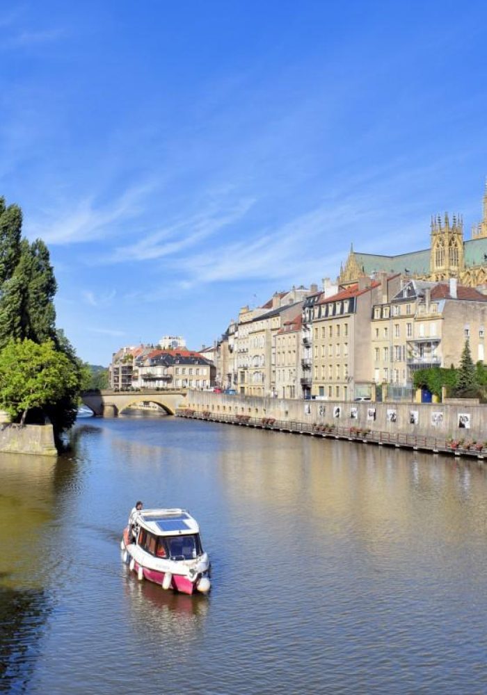 A small boat sails along a calm river lined with historic buildings and a church with a high spire, under a clear blue sky. Trees line the bank and a stone bridge can be seen in the background.