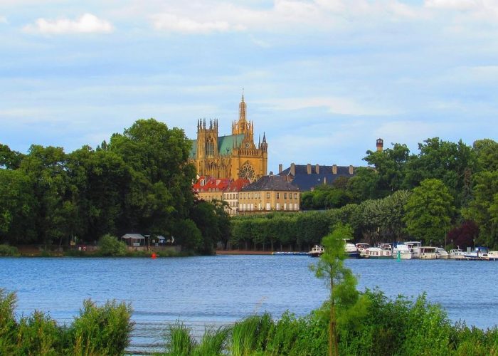 A large cathedral with soaring spires rises above the trees near one shore, with boats moored alongside and a clear blue sky overhead.