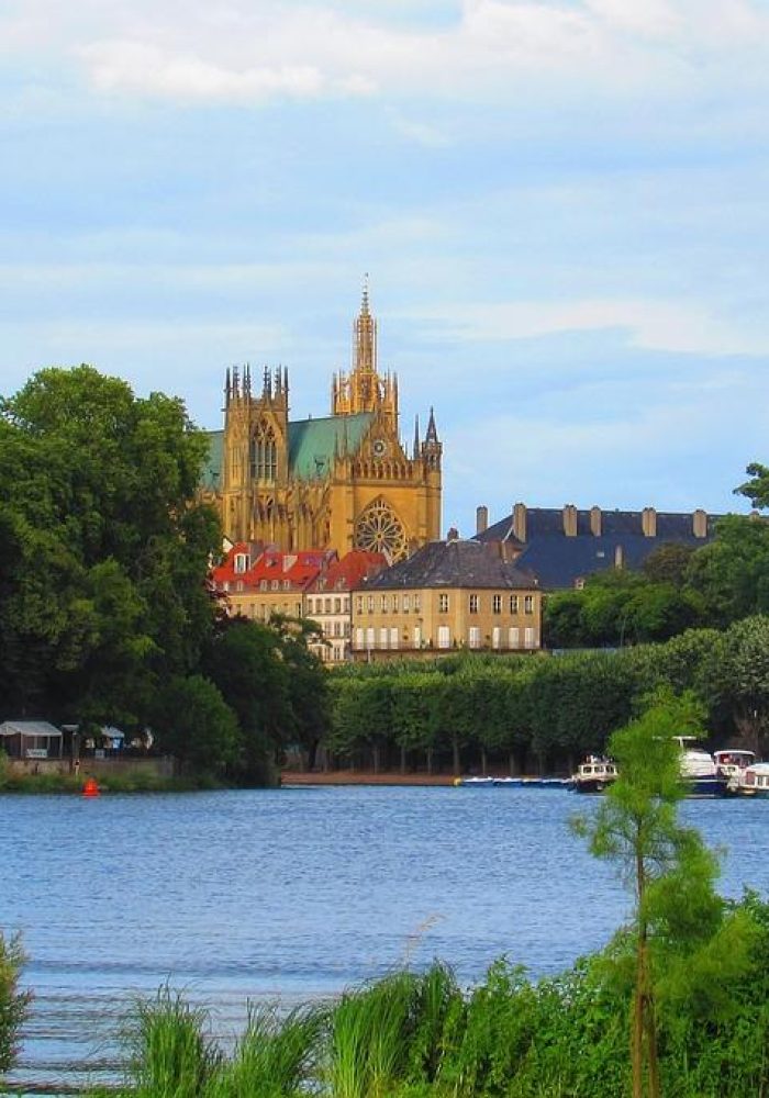A large cathedral with soaring spires rises above the trees near one shore, with boats moored alongside and a clear blue sky overhead.