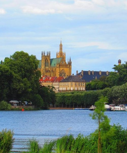 Une grande cathédrale aux flèches élevées s'élève au-dessus des arbres près d'une rive, avec des bateaux amarrés le long de la rive et un ciel bleu clair au-dessus de la tête.