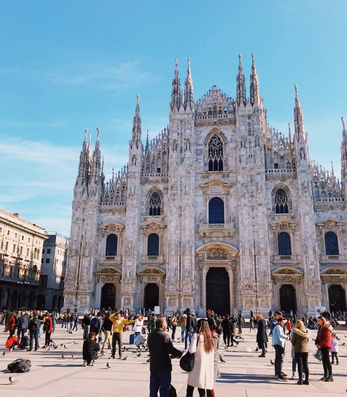 Les gens se rassemblent et prennent des photos devant la cathédrale de Milan (Duomo di Milano), une grande église de style gothique avec des flèches et des statues ornées, sous un ciel bleu clair.