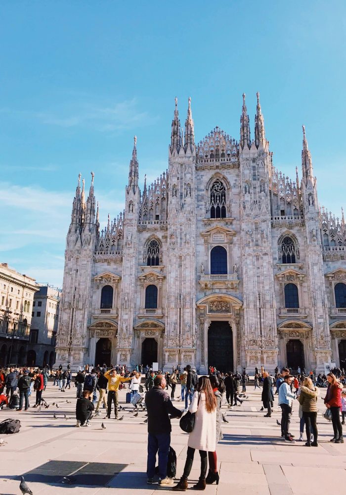 Les gens se rassemblent et prennent des photos devant la cathédrale de Milan (Duomo di Milano), une grande église de style gothique avec des flèches et des statues ornées, sous un ciel bleu clair.