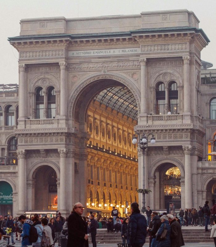 Une foule nombreuse se rassemble devant la grande entrée de la Galleria Vittorio Emanuele II à Milan, en Italie. L'arcade ornée est illuminée et flanquée de bâtiments historiques avec des boutiques et des devantures lumineuses.