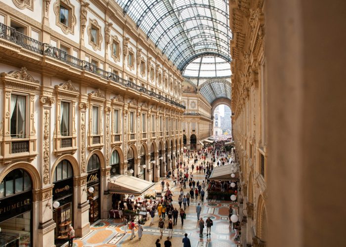 Une foule nombreuse traverse une galerie marchande ornée d'un toit en verre, à l'architecture élégante, aux fenêtres cintrées et aux sols carrelés de mosaïques, remplie de boutiques et de cafés.
