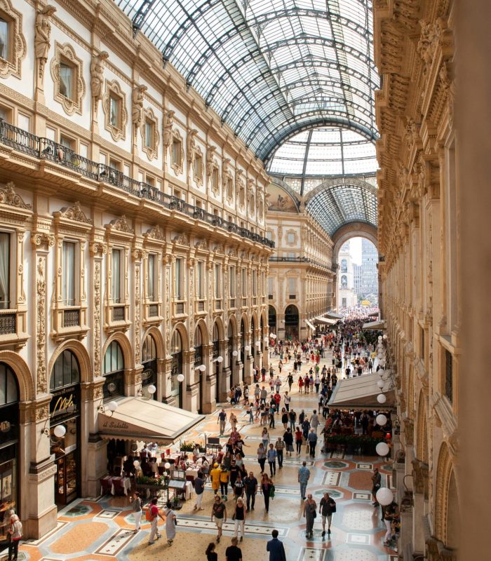 Une foule nombreuse traverse une galerie marchande ornée d'un toit en verre, à l'architecture élégante, aux fenêtres cintrées et aux sols carrelés de mosaïques, remplie de boutiques et de cafés.