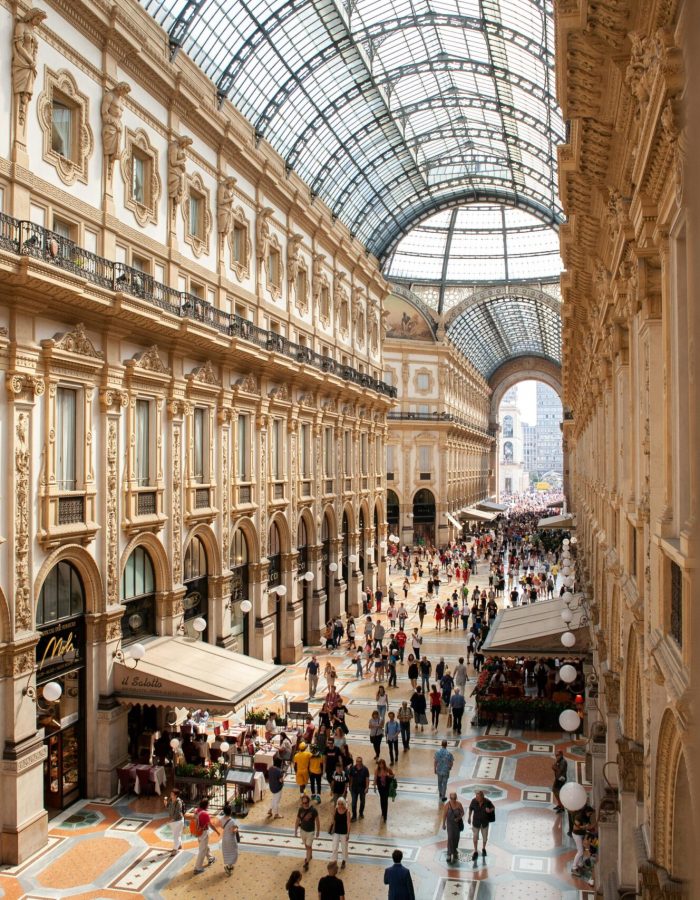 Une foule nombreuse traverse une galerie marchande ornée d'un toit en verre, à l'architecture élégante, aux fenêtres cintrées et aux sols carrelés de mosaïques, remplie de boutiques et de cafés.