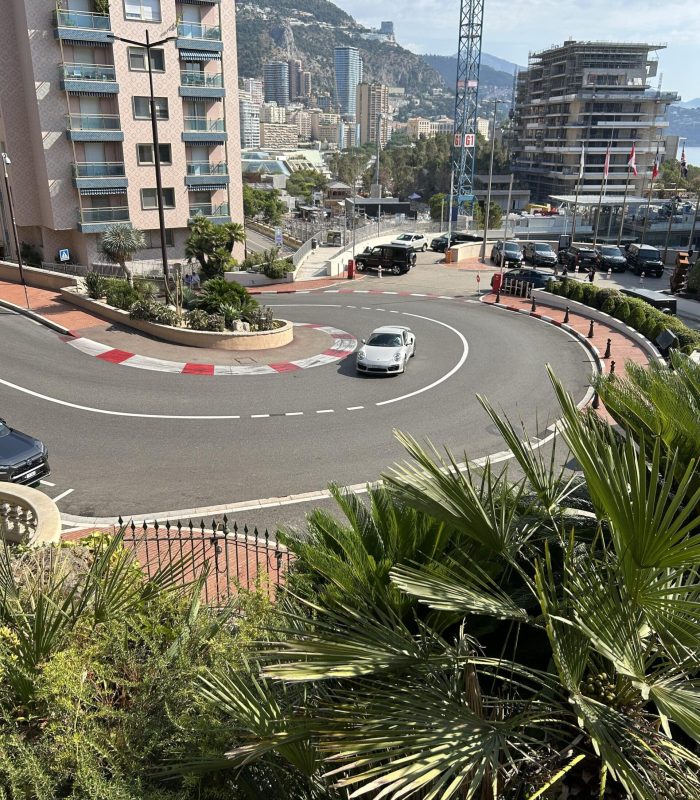 A white sports car negotiates a sharp bend in a city street surrounded by buildings and palm trees, with a mountainous background and partly cloudy sky.