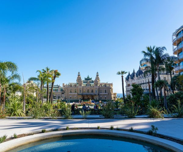 Une fontaine ronde au premier plan avec une végétation luxuriante et des palmiers, menant à un bâtiment historique orné sous un ciel bleu clair, flanqué de bâtiments modernes et classiques.