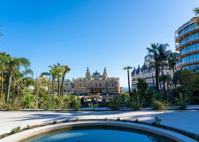 A round fountain in the foreground with lush vegetation and palm trees, leading to an ornate historic building under a clear blue sky, flanked by modern and classical buildings.