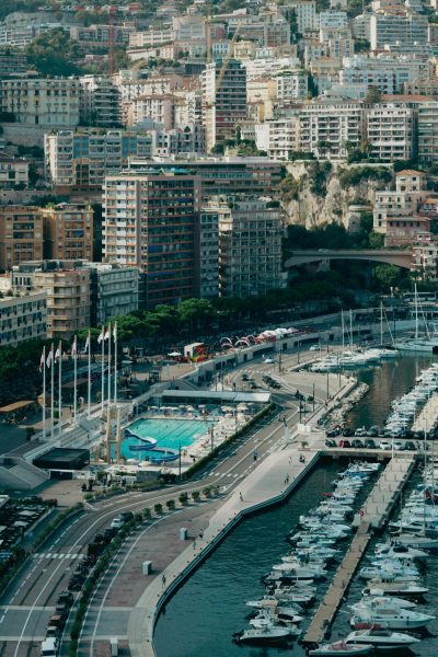 Vue aérienne d'un port de plaisance avec de nombreux yachts amarrés, une piscine entourée de drapeaux et, en arrière-plan, de grands immeubles densément peuplés qui s'élèvent à flanc de colline.