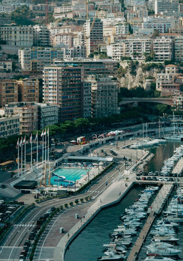 Aerial view of a marina with numerous yachts moored, a swimming pool surrounded by flags and, in the background, tall, densely-populated buildings rising up the hillside.