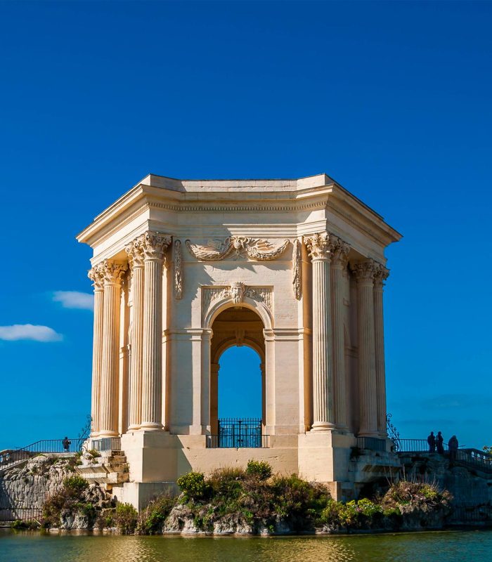 Un pavillon néoclassique en pierre avec des colonnes se trouve à côté d'un étang sous un ciel bleu clair, avec quelques personnes visibles près de la structure et des arbres à l'arrière-plan.