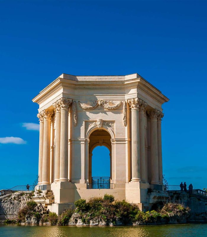 A neoclassical stone pavilion with columns stands beside a pond under a clear blue sky, with a few people visible near the structure and trees in the background.