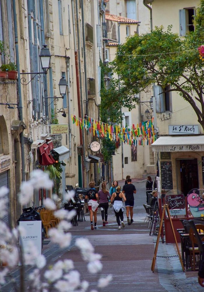 A narrow European street lined with old buildings, outdoor café tables and colourful flags hanging above. Five people walk along the cobbled path, while flowers appear in the foreground.
