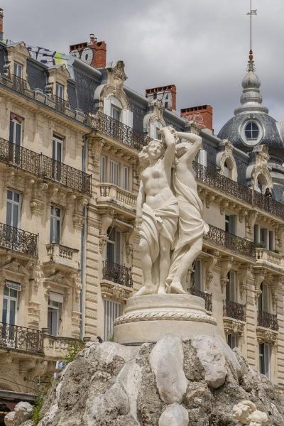 Une statue de pierre blanche représentant deux personnages se dresse sur un socle rocheux devant des bâtiments historiques ornés de balcons en fer forgé et de toits en dôme, sous un ciel nuageux.
