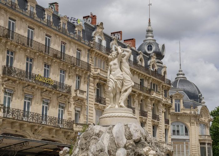 A white stone statue of two figures stands on a rocky plinth in front of historic buildings adorned with wrought-iron balconies and domed roofs, under a cloudy sky.