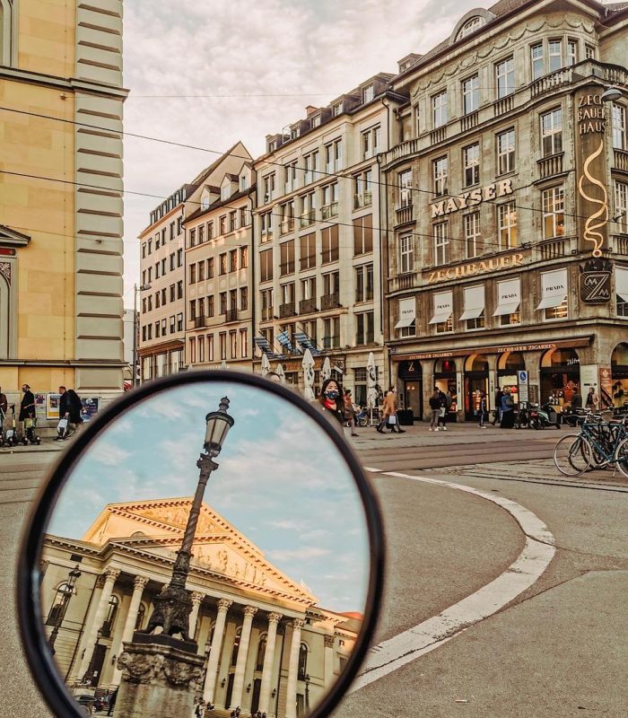 Scène de rue en ville avec des bâtiments historiques et des promeneurs ; au premier plan, un miroir rond reflète l'image d'un grand bâtiment à colonnes avec un fronton triangulaire, sous un ciel lumineux et partiellement nuageux.
