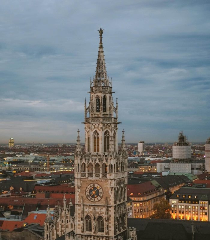 A tall, ornate clock tower of Gothic architecture rises above a cityscape at dusk, with roofs and buildings lit up beneath a cloudy sky.