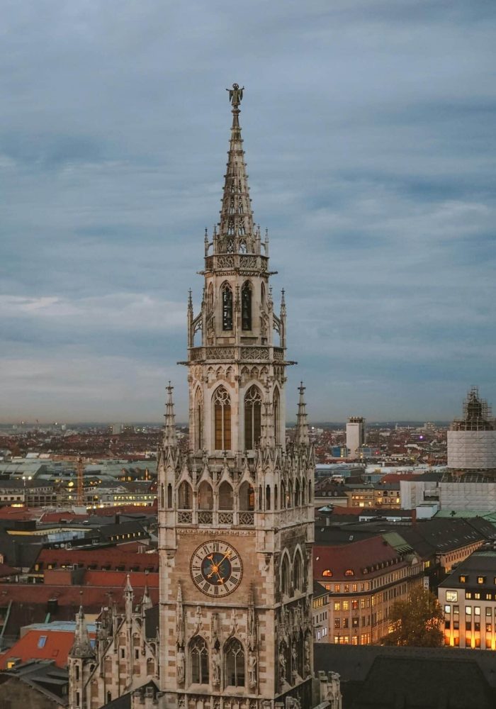 A tall, ornate clock tower of Gothic architecture rises above a cityscape at dusk, with roofs and buildings lit up beneath a cloudy sky.