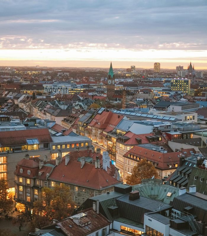 Vue aérienne d'une ville au coucher du soleil avec un mélange de bâtiments modernes et historiques, des toits rouges, des lampadaires lumineux et une église avec un clocher vert au centre.